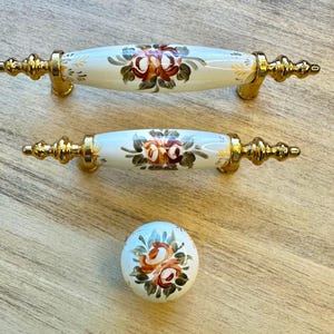 Op de afbeelding: Set van drie keramische ladegrepen met gouden accenten en bloemmotieven. De grepen hebben een witte achtergrond met oranje, gele en groene bloemen.
