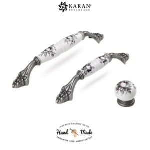 Könnte beinhalten: Zwei weiße Keramik-Schrankgriffe mit schwarzem Blumendesign und antikem Silberfinish. Ein passender Knopf ist ebenfalls abgebildet. Der Text "Hand Made" ist im Bild sichtbar.