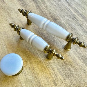 Pode incluir: Dois botões de armário de cerâmica branca com detalhes em metal dourado. Os botões têm uma forma arredondada, ligeiramente cónica.