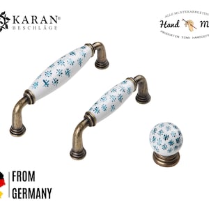 Könnte beinhalten: Schrankbeschlagsatz mit zwei Griffen und einem Knauf. Die Griffe und der Knauf haben eine weiße Keramikoberfläche mit blauem Blumenmuster und antiken Messingfarbenen Metallakzenten. Der Text "Hand Made" ist sichtbar.