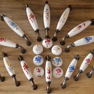 Könnte beinhalten: Ein Set mit 18 Keramik-Schrankknöpfen und -griffen mit Blumendesigns in verschiedenen Farben. Die Knöpfe und Griffe haben eine weiße Basis mit goldenen Akzenten. Die Knöpfe sind rund und die Griffe oval.