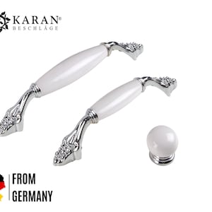 Könnte beinhalten: Zwei weiße Keramik-Schrankknöpfe mit silberner Oberfläche und verzierten Details. Die Knöpfe sind tropfenförmig mit abgerundeter Oberseite und spitzer Unterseite. Die Knöpfe werden vor einem weißen Hintergrund gezeigt.