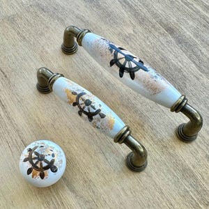 Pode incluir: Conjunto de três botões de armário de cerâmica com tema náutico. Os botões são brancos com um design de roda de navio em ouro e preto. Os botões têm um acabamento em latão.