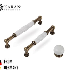 Könnte beinhalten: Zwei Griffe im antiken Stil und ein Knopf. Die Griffe haben eine weiße Keramikmitte und bronzefarbene Metallenden. Der Knopf ist aus weißer Keramik mit einem bronzefarbenen Sockel. Der Text "KARAN BESCHLÄGE" ist sichtbar. "FROM GERMANY" ist ebenfalls sichtbar.