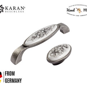 Peut inclure: Une paire de boutons de cabinet en argent antique avec des inserts en céramique blanche ornés d'un motif floral. Les boutons sont de forme ovale et ont une poignée légèrement incurvée. Le texte "Hand Made" est visible en arrière-plan.