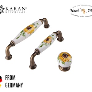 Op de afbeelding: Set van drie keramische kastknoppen met zonnebloemontwerpen. De knoppen zijn wit met gele en groene zonnebloemen. De knoppen hebben een messing afwerking.