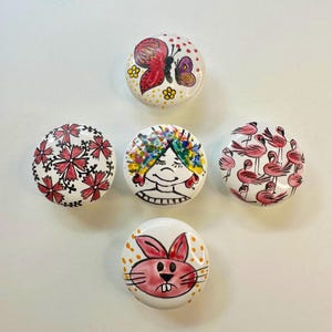 May include: Five round ceramic knobs with different painted designs. One knob features a red butterfly, another has a pink floral pattern, a third has a woman with colorful hair, a fourth has a pink bunny, and the last has a group of pink flamingos.