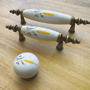 Può includere: Due pomelli per armadi in ceramica bianca con dettagli dorati e un design a forma di grano. I pomelli sono su una superficie in legno. C'è anche un singolo pomello in ceramica bianca con un design a forma di grano su una superficie in legno.