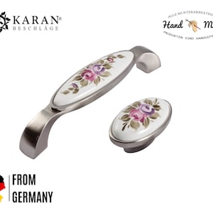 Può includere: Una coppia di pomelli per armadi in argento con un inserto ovale in ceramica bianca con un motivo floreale dipinto a mano in tonalità rosa e viola. Il testo "Hand Made" è visibile sullo sfondo.