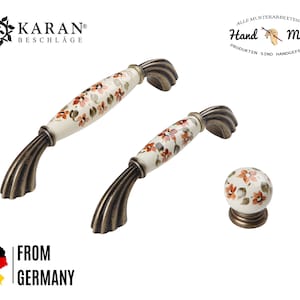 Op de afbeelding: Twee antieke bronzen kastknoppen met bloemmotieven. De knoppen zijn gemaakt van keramiek met een witte basis en oranje bloem accenten. De knoppen zijn handgeschilderd en hebben een vintage uitstraling.