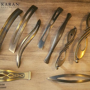 Puede incluir: Surtido de ocho tiradores de puerta de armario de oro antiguo con varios diseños. Los tiradores están dispuestos sobre una superficie de madera. KARAN BESCHLAGE está impreso en la esquina superior izquierda de la imagen.