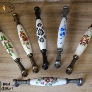 Op de afbeelding: Zes keramische ladegrepen met verschillende bloemenontwerpen. De grepen zijn wit met verschillende gekleurde bloemenpatronen. De grepen hebben een metalen basis in antiek messing en zilveren afwerking. De tekst "Hand Made" is zichtbaar in de linkerbovenhoek van de afbeelding.