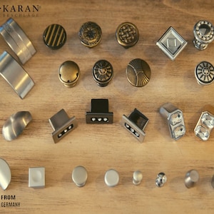 Può includere: Assortimento di 20 pomelli e maniglie per armadi in vari stili e finiture, tra cui argento, oro, nero e cristallo. I pomelli e le maniglie sono disposti su una superficie in legno. KARAN® BESCHLAGE FROM GERMANY è stampato sulla superficie.