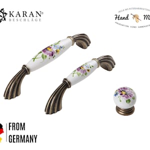 Könnte beinhalten: Zwei antike Messing-Schrankknöpfe mit weißen Keramiksockeln, die mit floralen Designs verziert sind. Die Knöpfe sind handbemalt mit lila, gelben und blauen Blumen. Der Text "Hand Made" ist im Hintergrund sichtbar.