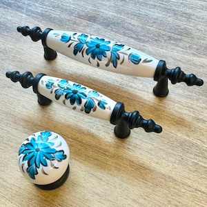 Könnte beinhalten: Set aus drei Keramik-Schubladengriffen mit weißem Hintergrund und blauem Blumendesign. Die Griffe haben schwarze Metallsockel.