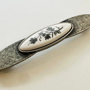 Pode incluir: Uma puxador de armário de tom prateado com um inserto oval de cerâmica branca com um design floral preto.