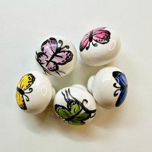 Può includere: Cinque pomelli per cassetti in ceramica bianca con disegni di farfalle dipinte. Le farfalle sono di vari colori, tra cui rosa, giallo, blu e verde.
