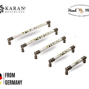 Könnte beinhalten: Fünf weiße Keramik-Schrankgriffe mit einem grünen Olivenzweig-Design. Die Griffe haben eine gebürstete Messing-Oberfläche und sind in absteigender Größenordnung angeordnet.