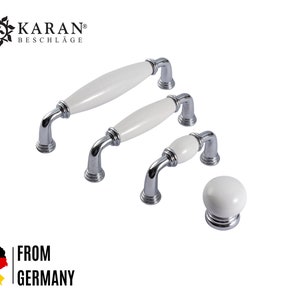 Op de afbeelding: Set van drie witte keramische kastknoppen met chroomaccenten. De knoppen zijn in verschillende maten en zijn gerangschikt in een rij van groot naar klein. De kleinste knop is een ronde knop. De tekst "KARAN® BESCHLAGE" is zichtbaar in de linkerbovenhoek van de afbeelding.