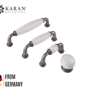 Könnte beinhalten: Drei weiße Keramik-Schrankknöpfe mit antiken silbernen Metallsockeln. Die Knöpfe sind in verschiedenen Größen. Der größte Knopf ist 10 cm lang, der mittlere Knopf ist 7,5 cm lang und der kleinste Knopf ist 5 cm lang. Die Knöpfe werden in Deutschland hergestellt.