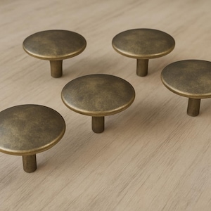 May include: Five antique brass cabinet knobs with a round, flat top and a short, cylindrical stem. The knobs are arranged on a light-colored wooden surface. The knobs have a slightly aged, vintage appearance.