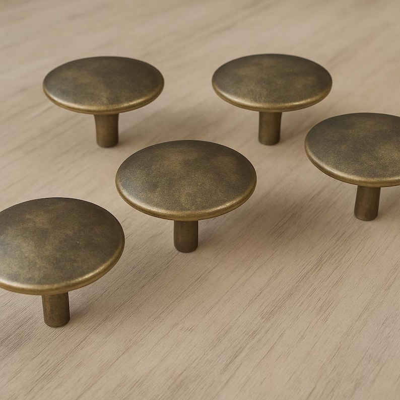 May include: Five antique brass cabinet knobs with a round, flat top and a short, cylindrical stem. The knobs are arranged on a light-colored wooden surface. The knobs have a slightly aged, vintage appearance.