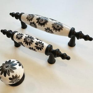 Puede incluir: Tres tiradores de cajón de cerámica blanca y negra con diseños florales. Los tiradores tienen una base de metal negro y un cuerpo de cerámica blanca con patrones florales negros. Los tiradores son de varios tamaños.