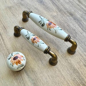 Pode incluir: Dois botões de armário de cerâmica branca com um design floral e detalhes em metal dourado. Também é mostrado um botão menor correspondente.
