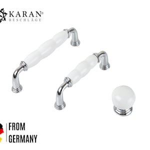Pode incluir: Dois botões de armário de cerâmica branca com detalhes cromados e um único botão de cerâmica branca com base cromada.  KARAN® BESCHLAGE está impresso na imagem.