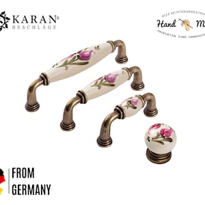 Pode incluir: Conjunto de três botões e puxadores de armário de cerâmica com um design floral em tons de rosa e branco. Os botões e puxadores têm um acabamento em latão escovado. O texto "Hand Made" é visível no fundo.