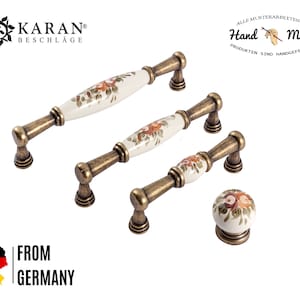 Peut inclure: Ensemble de trois boutons de cabinet en or antique avec des poignées en céramique blanche ornées de motifs floraux. Les boutons sont peints à la main et ont un aspect vintage.  "Hand Made" et "From Germany" sont imprimés sur l'image.