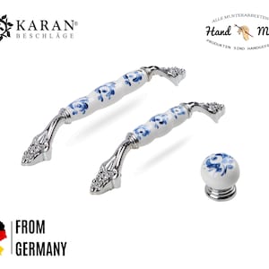 Könnte beinhalten: Zwei weiße Keramik-Schrankknöpfe mit blauen Blumenmustern und silbernen Akzenten. Die Knöpfe werden vor einem weißen Hintergrund gezeigt. Der Text "Hand Made" ist in der oberen rechten Ecke des Bildes sichtbar.