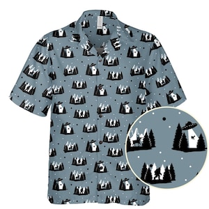 May include: A short-sleeved button-down shirt with a light blue background and a repeating pattern of black and white illustrations. The design includes trees, UFOs, and Bigfoot-like figures. The shirt has a classic collar and button closure.