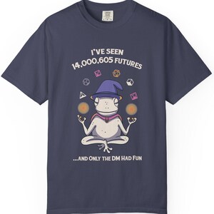 May include: Navy blue t-shirt with a cartoon frog wearing a wizard hat and holding orbs. Text reads "I'VE SEEN 14,000,605 FUTURES ...AND ONLY THE DM HAD FUN". Various geometric shapes are also present.