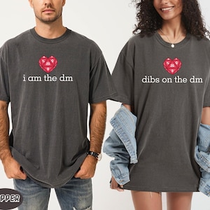 Puede incluir: Dos camisetas gris oscuro con los textos "i am the dm" y "dibs on the dm". Cada camiseta tiene un gráfico de dado rojo en forma de corazón. Las camisetas son de manga corta y tienen un aspecto vintage.