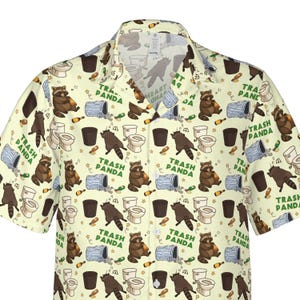 Pode incluir: Uma camisa de manga curta com bot&otilde;es em cor creme claro, apresentando uma estampa de guaxinins castanhos, sanitas brancas, caixotes do lixo azuis e garrafas castanhas. O texto "TRASH PANDA" est&aacute; impresso em verde por todo o padr&atilde;o.