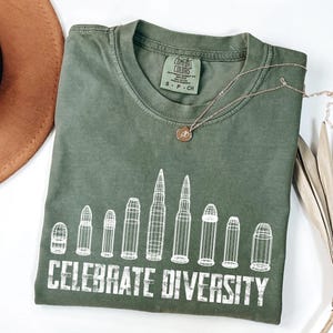 Peut inclure: T-shirt vert olive avec l'inscription blanche "CELEBRATE DIVERSITY". Le t-shirt présente un motif de plusieurs balles. Un collier en or avec un petit pendentif est posé sur le t-shirt.