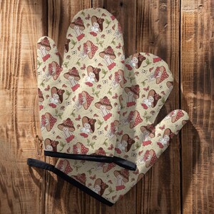 May include: A pair of oven mitts with a whimsical design featuring a repeating pattern of mushrooms and bunnies wearing hats. The mitts are beige with brown trim and have a black hanging loop.