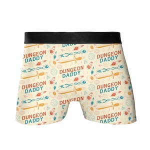 May include: White boxer shorts with a black waistband and a repeating pattern of swords, dice, potions, and the text "Dungeon Daddy".