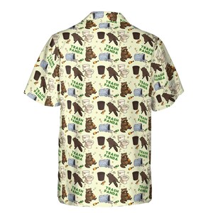 Pode incluir: Uma camisa de manga curta com bot&otilde;es em cor creme claro, apresentando um padr&atilde;o repetitivo de guaxinins, latas de lixo, vasos sanit&aacute;rios e garrafas. O texto "TRASH PANDA" est&aacute; impresso em verde entre pequenos &iacute;cones de estrelas.