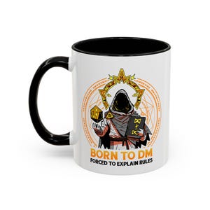 Könnte beinhalten: Weiße Keramik-Tasse mit schwarzem Henkel und Innenraum. Die Tasse zeigt eine Grafik einer vermummten Figur, die einen Würfel und ein Buch hält, mit dem Text "BORN TO DM FORCED TO EXPLAIN RULES" in Orange.