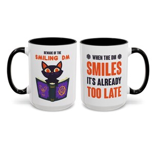May include: White ceramic mug with black handle and interior. One side features a black cat reading a book with the text "BEWARE OF THE SMILING DM". The other side says "WHEN THE DM SMILES IT'S ALREADY TOO LATE".