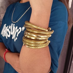 Könnte beinhalten: Ein Satz goldfarbener Metallarmreifen mit einem strukturierten Design. Die Armreifen sind auf dem Handgelenk einer Person gestapelt.