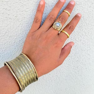 May include: A gold-toned bracelet with a woven design, worn on a person's wrist. The person is also wearing several gold rings on their fingers.