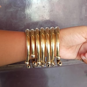 May include: A set of eight gold-colored metal bangles stacked on a person's wrist. The bangles are smooth and have a rounded shape.