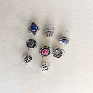 May include: Assortment of nine silver nose rings with various designs, including a flower, a star, and a geometric shape. Some rings feature blue and red gemstones.