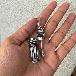 Peut inclure: Un collier avec un pendentif en argent composé d'un charme cylindrique qui s'ouvre et d'un plus petit charme décoratif attaché au sommet. Les charmes sont finement détaillés avec des motifs géométriques.