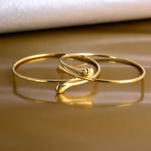 Könnte beinhalten: Goldfarbene Metallarmreifen. Ein Armreif hat ein einzigartiges Design mit tropfenförmigen Akzenten. Der andere Armreif ist ein schlichtes, klassisches Design. Der Schmuck wird auf einer reflektierenden, goldfarbenen Oberfläche präsentiert.
