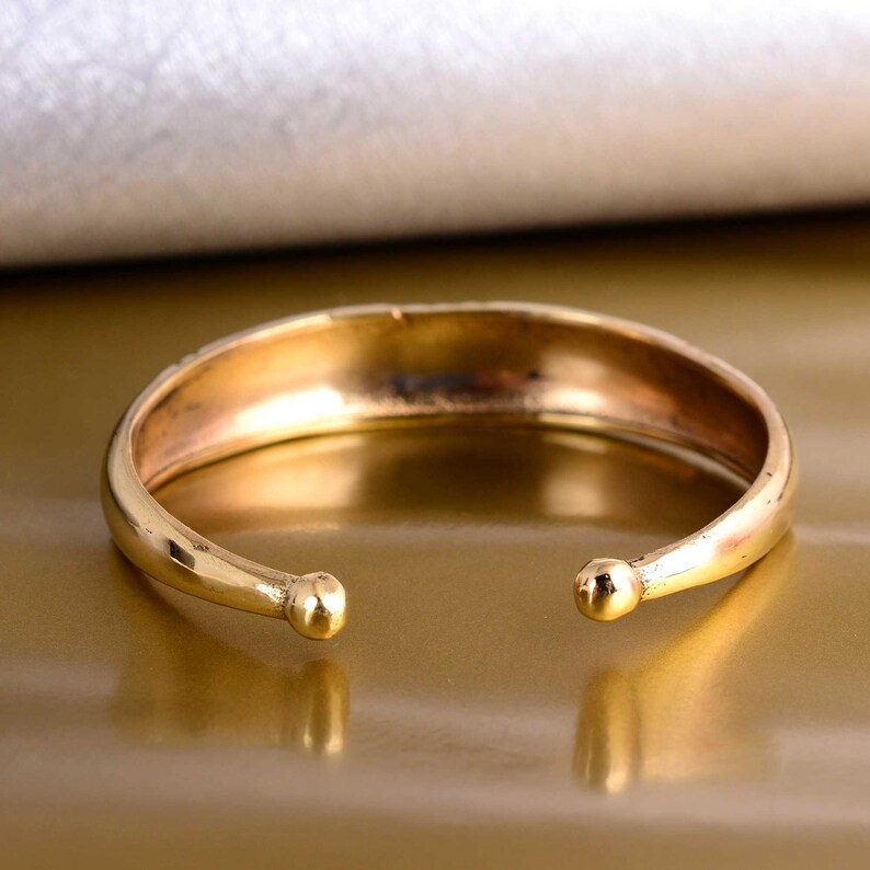 Może przedstawiać: Złota bransoletka typu bangle z zaokrąglonymi końcami. Bransoletka jest otwarta i ma gładkie, polerowane wykończenie.