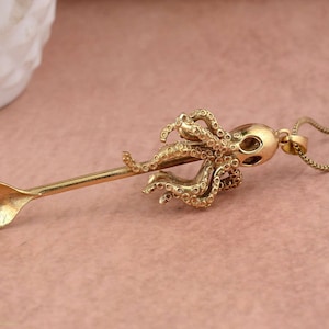 May include: A gold-toned necklace featuring an octopus pendant. The octopus is attached to a spoon-shaped charm.
